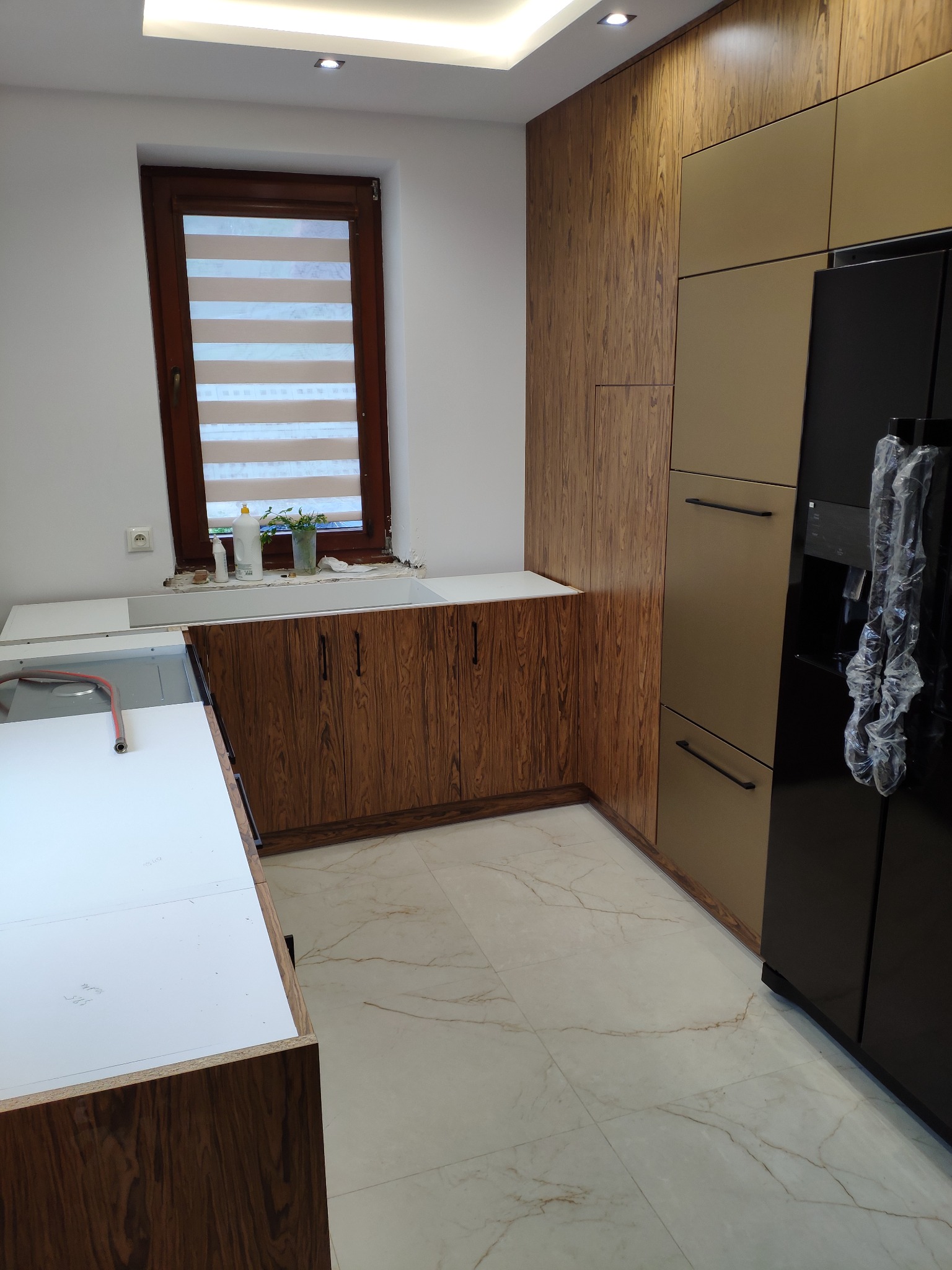 Montaż kuchni na wymiar: szafki dolne z fornirem w odcieniach brązu, biały blat roboczy w trakcie instalacji, obok wysoka zabudowa z szafkami w kolorze beżowym i czarna lodówka z dozownikiem wody...