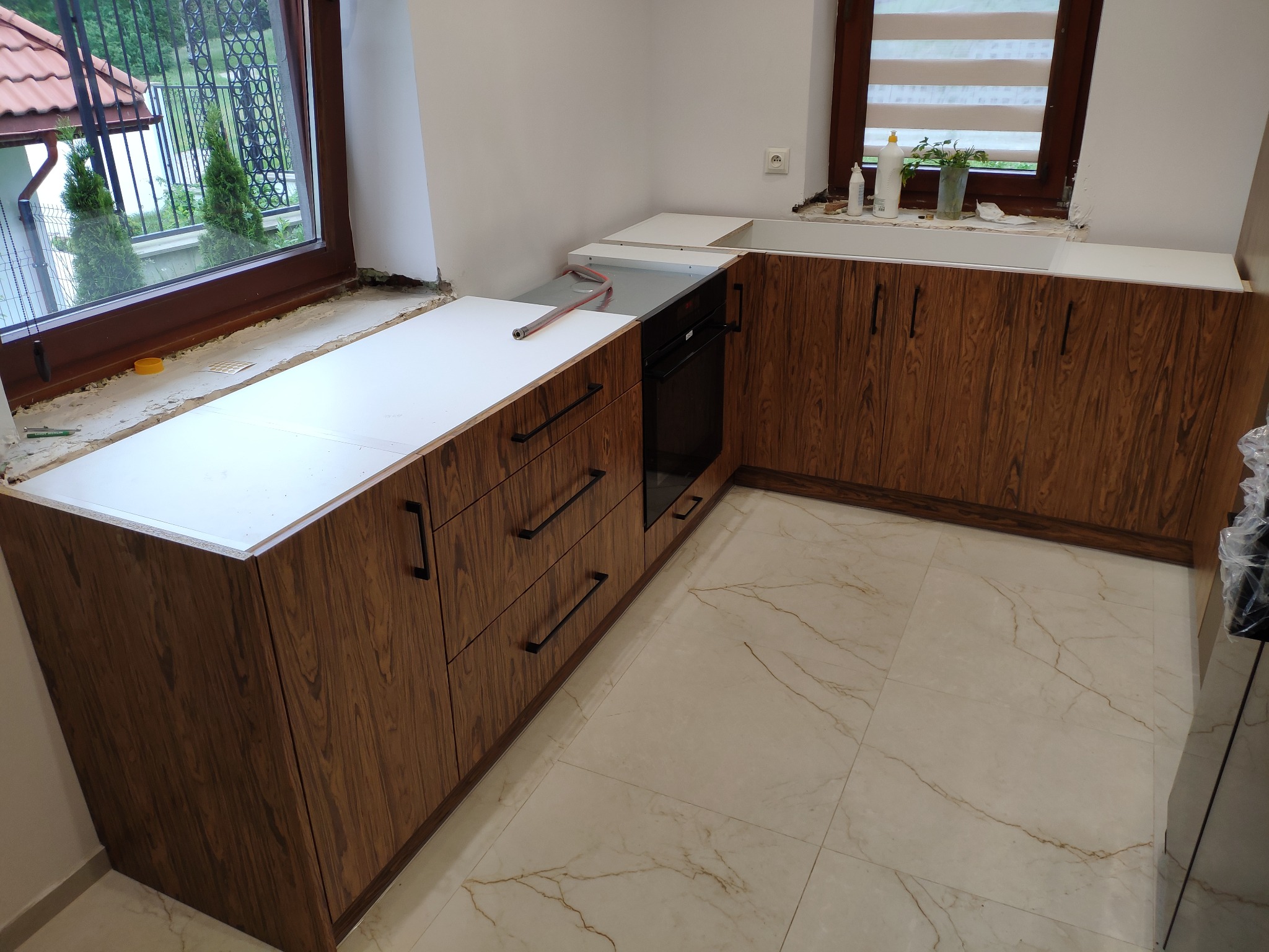 Fragment montażu kuchni: szafki dolne z frontami w odcieniach brązu z wyraźnym rysunkiem drewna, białe blaty, czarny piekarnik w zabudowie, widoczne okno z roletami i okno z widokiem na zieleń...