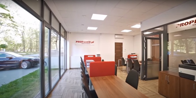 Jasne biuro agencji nieruchomości z dużymi oknami i logo 'PROPERCO.pl'. Widok na zewnątrz z zaparkowanym samochodem. Minimalistyczny wystrój, biurka i krzesła.