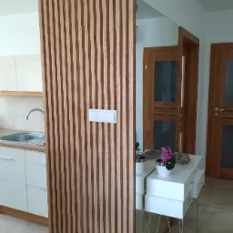 Drewniana ścianka działowa z pionowych listew oddzielająca kuchnię od przedpokoju, z widocznymi włącznikami światła i fragmentem lustra z białą konsolą i kwiatami.