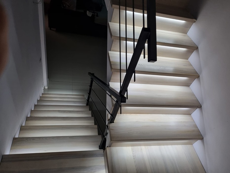 Nowoczesne schody z jasnego drewna z podświetleniem LED pod każdym stopniem i czarną, minimalistyczną poręczą.
