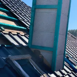 Montaż czarnej blachodachówki trapezowej wokół komina z szalunkiem w kolorze turkusowym, widoczne fragmenty membrany dachowej i drewniana łata.