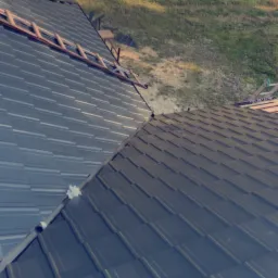 Metalowy dach z widoczną drabiną i elementami montażowymi, sfotografowany z góry na tle zieleni i drogi.