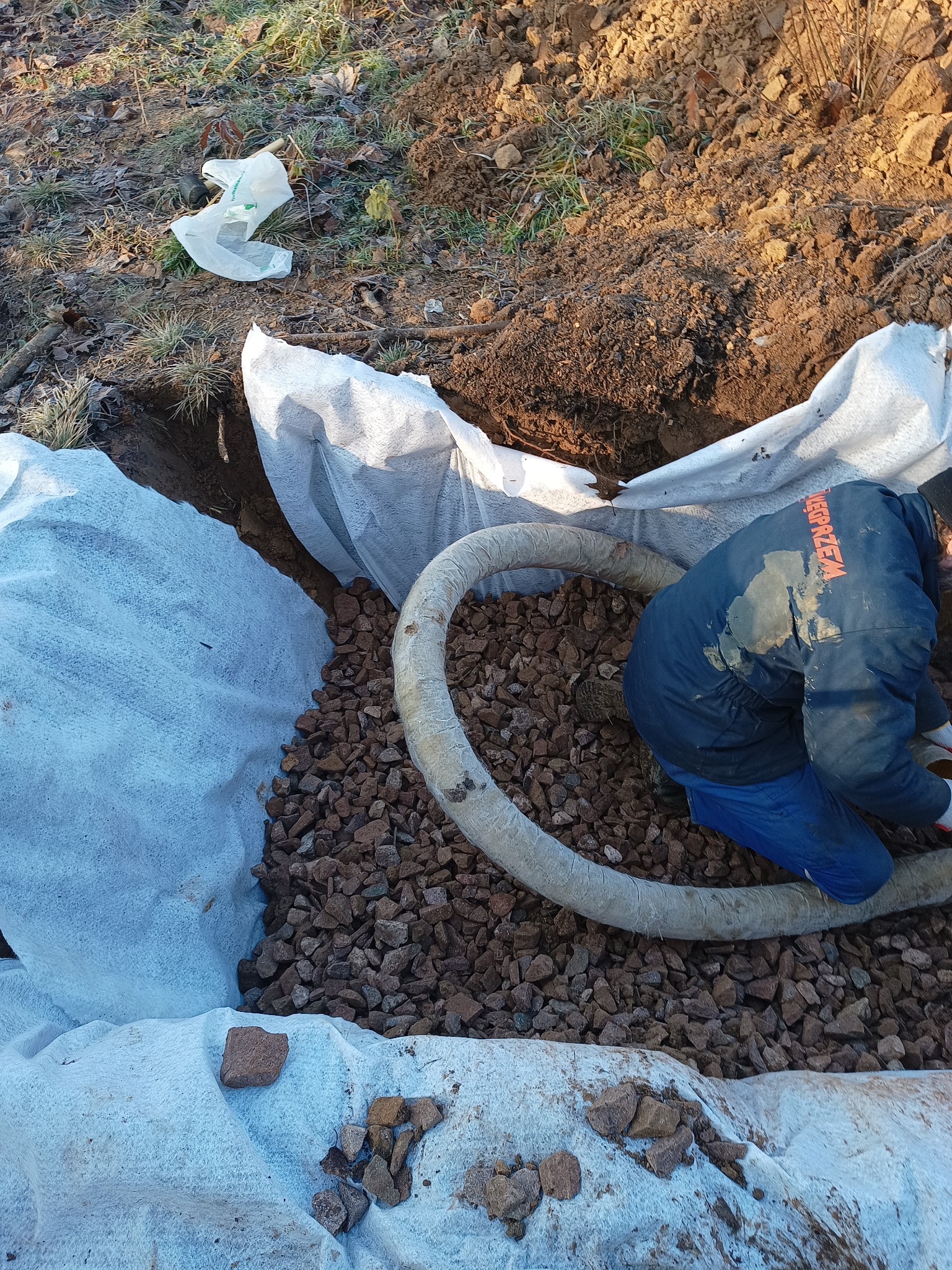 Pracownik w trakcie układania drenażu z rur i kamieni w wykopie wyłożonym geowłókniną, widoczna ziemia i resztki roślinności.