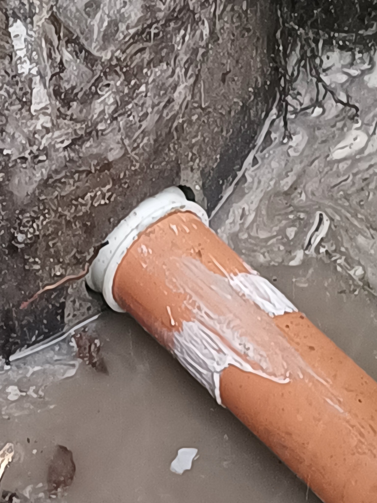 Pomarańczowa rura kanalizacyjna uszczelniona białą pastą w wykopie ziemnym, widoczne ślady wilgoci.