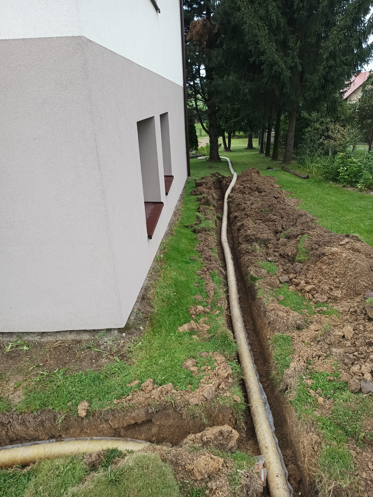 Rura drenarska ułożona w wykopie wzdłuż ściany budynku, widoczne świeżo wykopana ziemia i trawa.