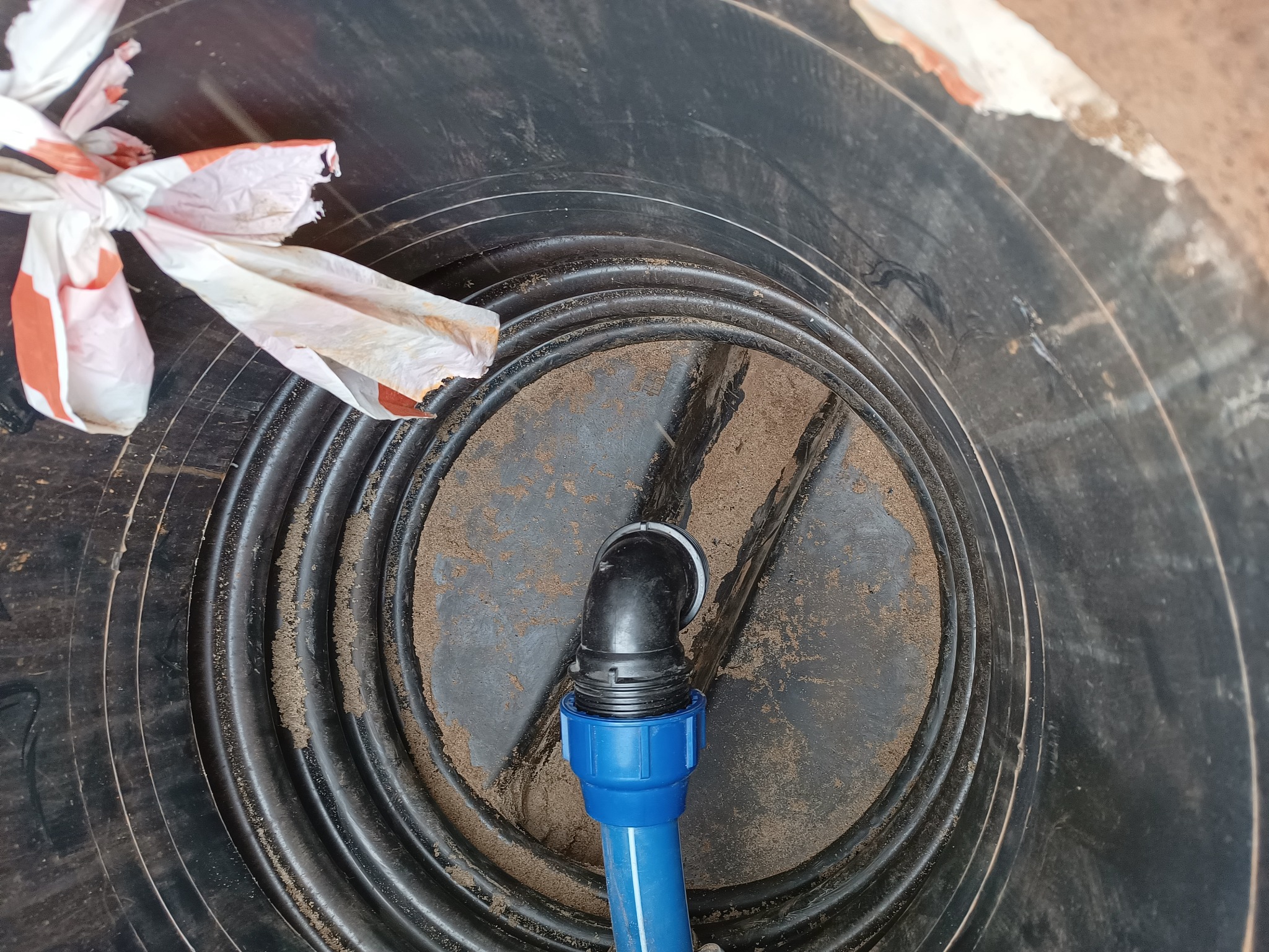 Wnętrze czarnego zbiornika z żebrowaną strukturą, z piaskiem na dnie i podłączonym niebieskim rurociągiem z czarnym kolankiem, obok leży częściowo rozerwana reklamówka.