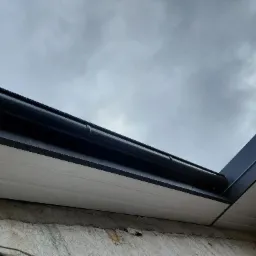 Nowe, czarne rynny zamontowane na białej podbitce dachowej, widok z dołu na tle pochmurnego nieba.