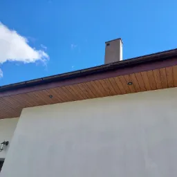 Fragment domu z brązowym podbitką dachową z oświetleniem punktowym, widoczne rynny, komin i fragment elewacji w jasnym odcieniu na tle błękitnego nieba z chmurami.