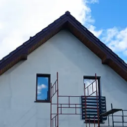 Ściana szczytowa domu z dwoma oknami, na tle błękitnego nieba z chmurami, częściowo zasłonięta rusztowaniem malarskim.