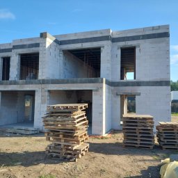 Eryk Konarski - Surowy stan dwukondygnacyjnego domu z betonu komórkowego w słoneczny dzień. Widoczne palety z materiałami budowlanymi przed budynkiem.