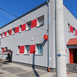 Eryk Konarski - Szary budynek z czerwonymi akcentami wokół okien i wejścia, widoczny z zewnątrz w słoneczny dzień. Na pierwszym planie słup i fragment rusztowania.