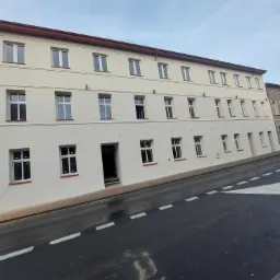 Odnowiona elewacja budynku wielorodzinnego w jasnych kolorach, z widoczną częścią rusztowania po lewej stronie, sfotografowana z poziomu ulicy.