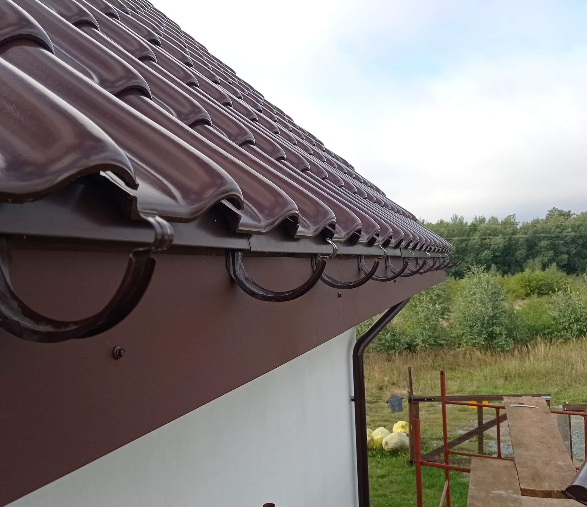 Fragment brązowego dachu z dachówką ceramiczną i rynnami w Nakle nad Notecią, widok z dołu, w tle zieleń i błękitne niebo.