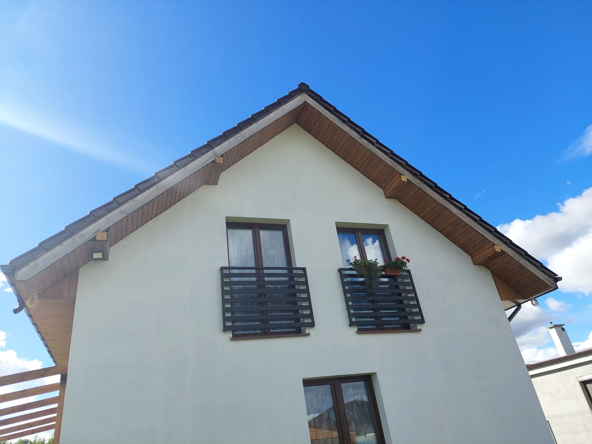 Nowoczesny dom murowany z białymi ścianami, ciemnym dachem i balkonami z kwiatami pod oknami, na tle błękitnego nieba z chmurami.