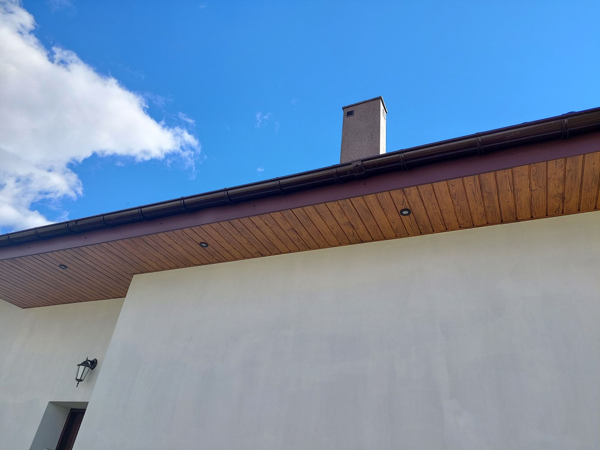 Fragment domu z brązowym podbitką dachową z oświetleniem punktowym, widoczne rynny, komin i fragment elewacji w jasnym odcieniu na tle błękitnego nieba z chmurami.