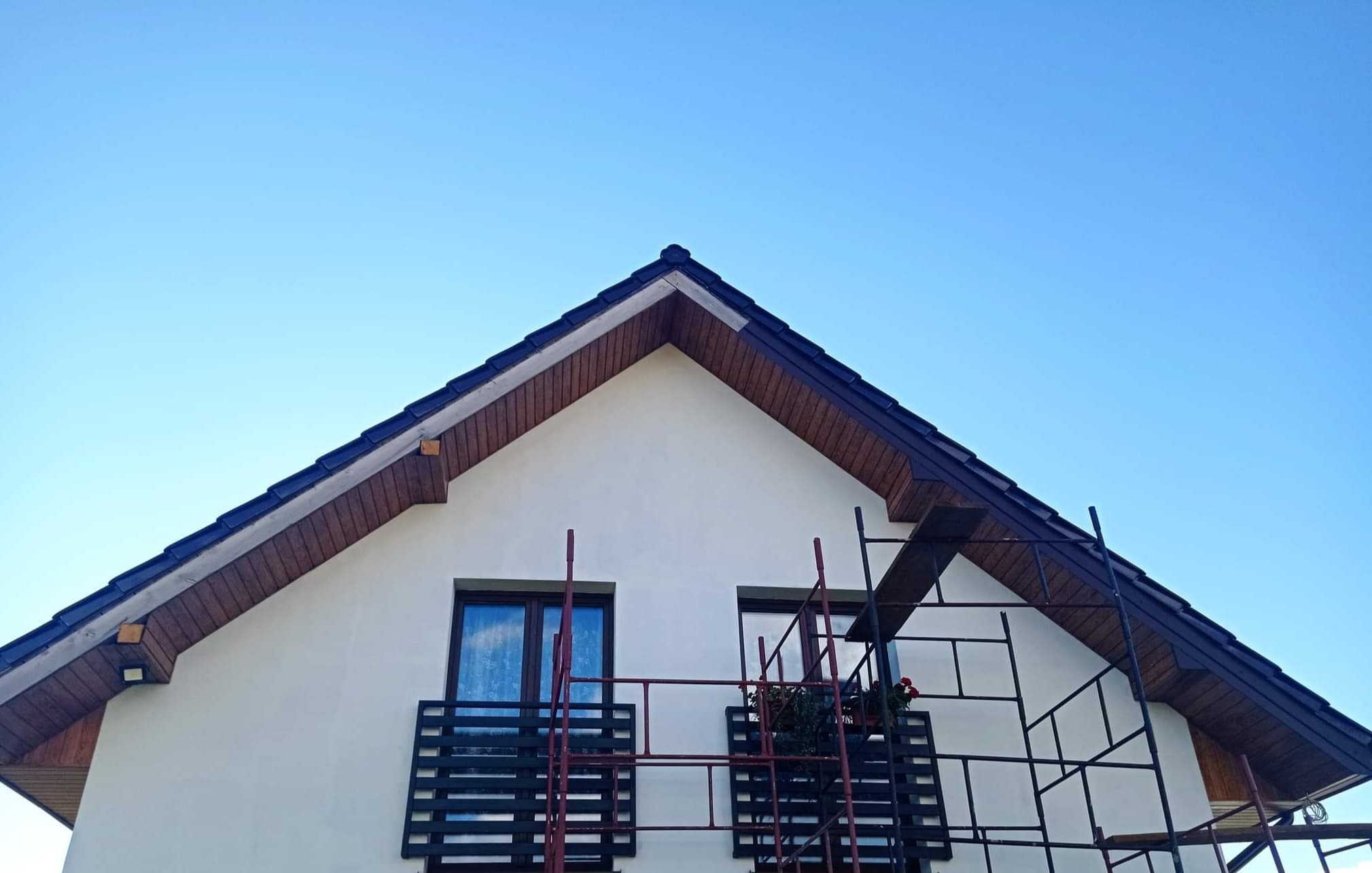 Fragment domu z ciemnym dachem i jasną elewacją, widoczne rusztowanie przed balkonem z kwiatami, błękitne niebo w tle.
