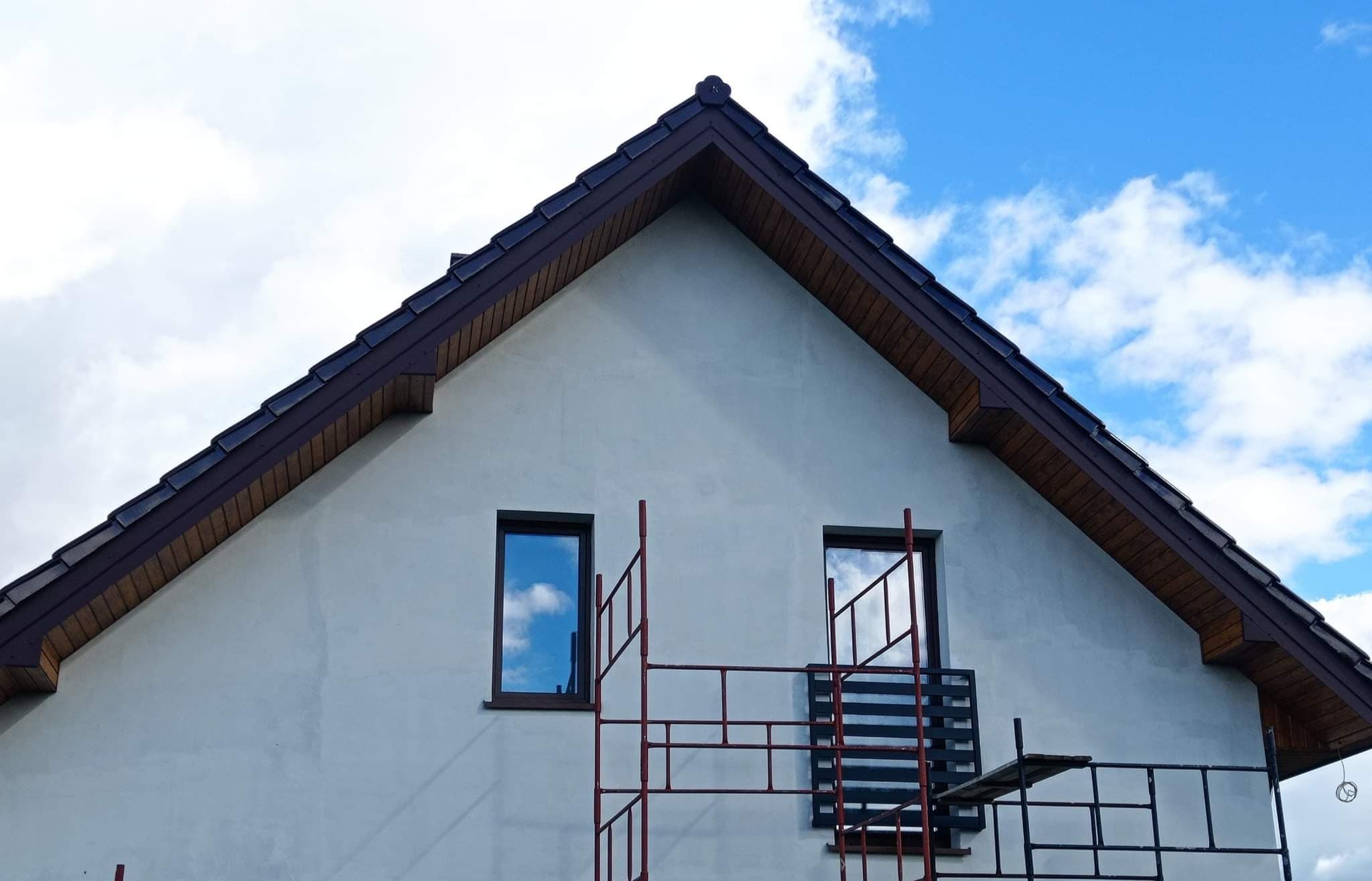 Ściana szczytowa domu z dwoma oknami, na tle błękitnego nieba z chmurami, częściowo zasłonięta rusztowaniem malarskim.