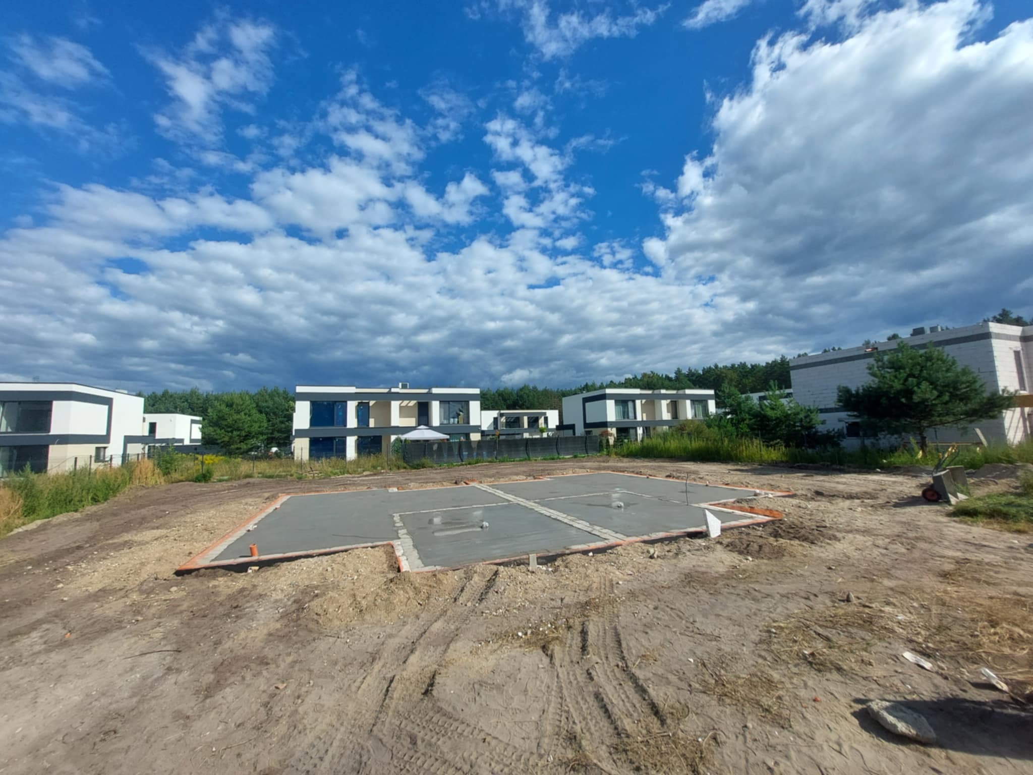 Wylana płyta fundamentowa pod budowę nowoczesnego domu jednorodzinnego na działce z widokiem na las i błękitne niebo z chmurami.