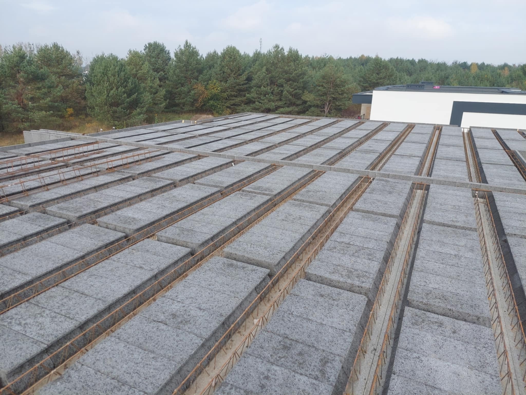 Szary strop Teriva zbrojony stalą na budowie domu jednorodzinnego, widok z góry. W tle zieleń drzew i fragment budynku.