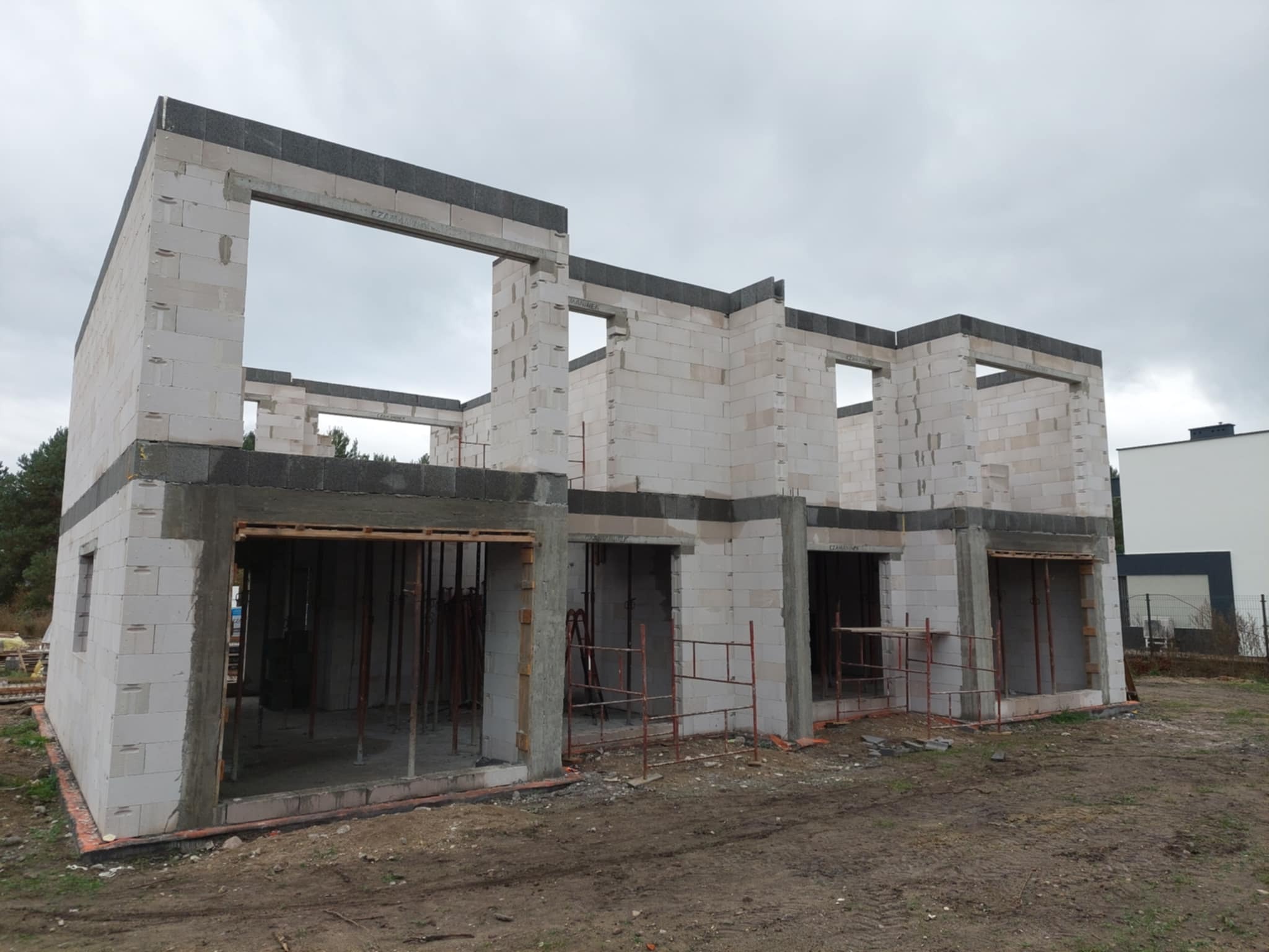 Surowy stan domu z betonu komórkowego w trakcie budowy. Widoczne ściany, otwory okienne i drzwiowe, szalunki oraz podpory. Tło stanowi pochmurne niebo i fragment zieleni.