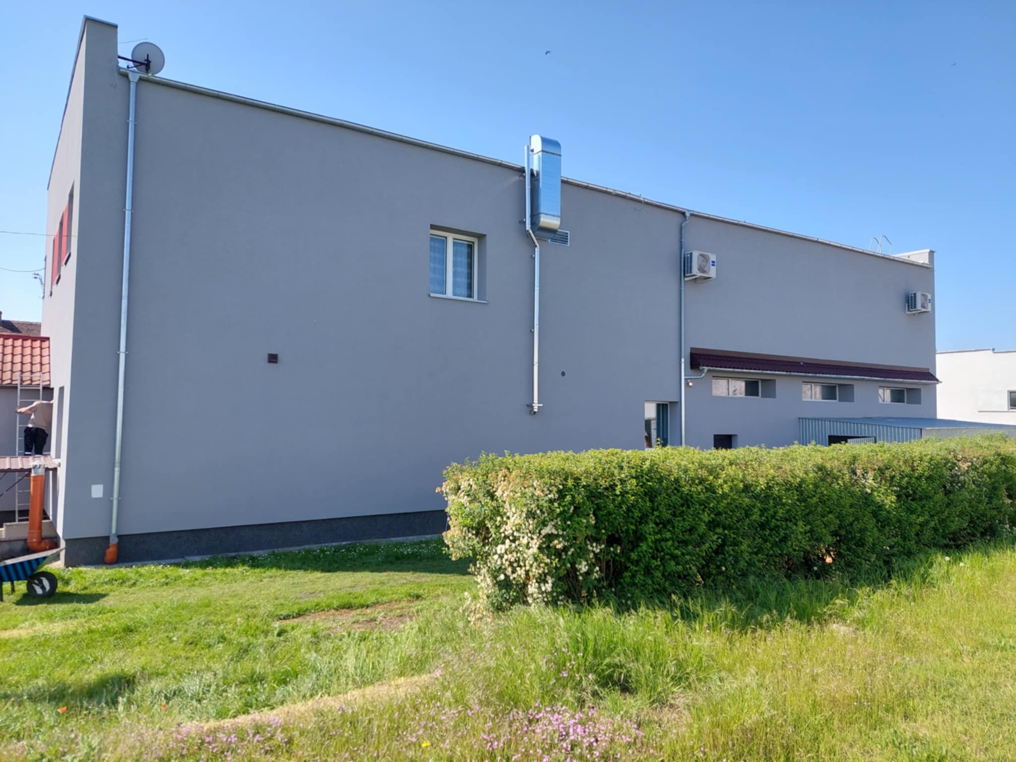 Szary budynek z blachy trapezowej z systemem wentylacyjnym na elewacji, widok z poziomu trawnika. Na rusztowaniu stoi osoba.