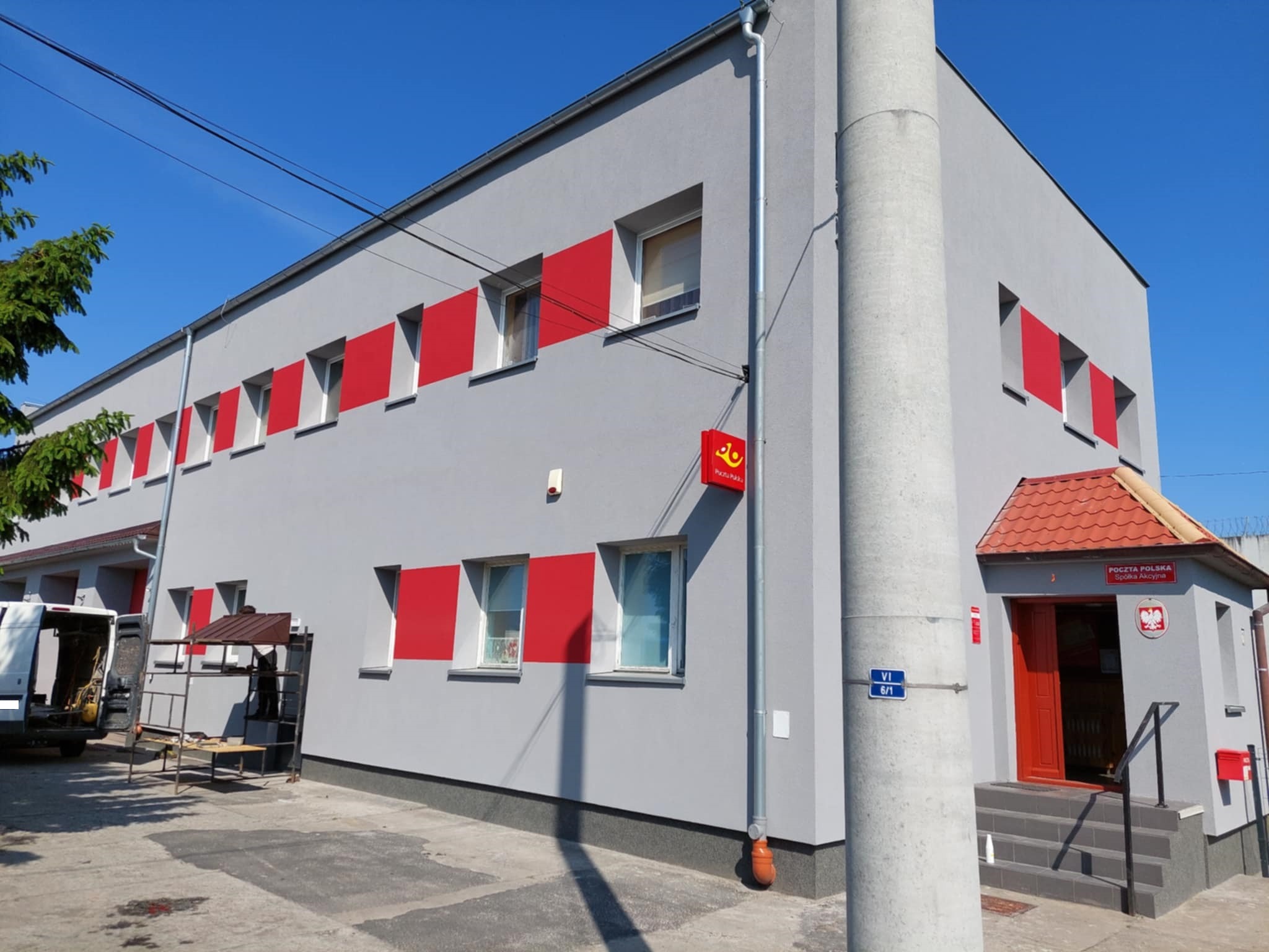 Szary budynek z czerwonymi akcentami wokół okien i wejścia, widoczny z zewnątrz w słoneczny dzień. Na pierwszym planie słup i fragment rusztowania.