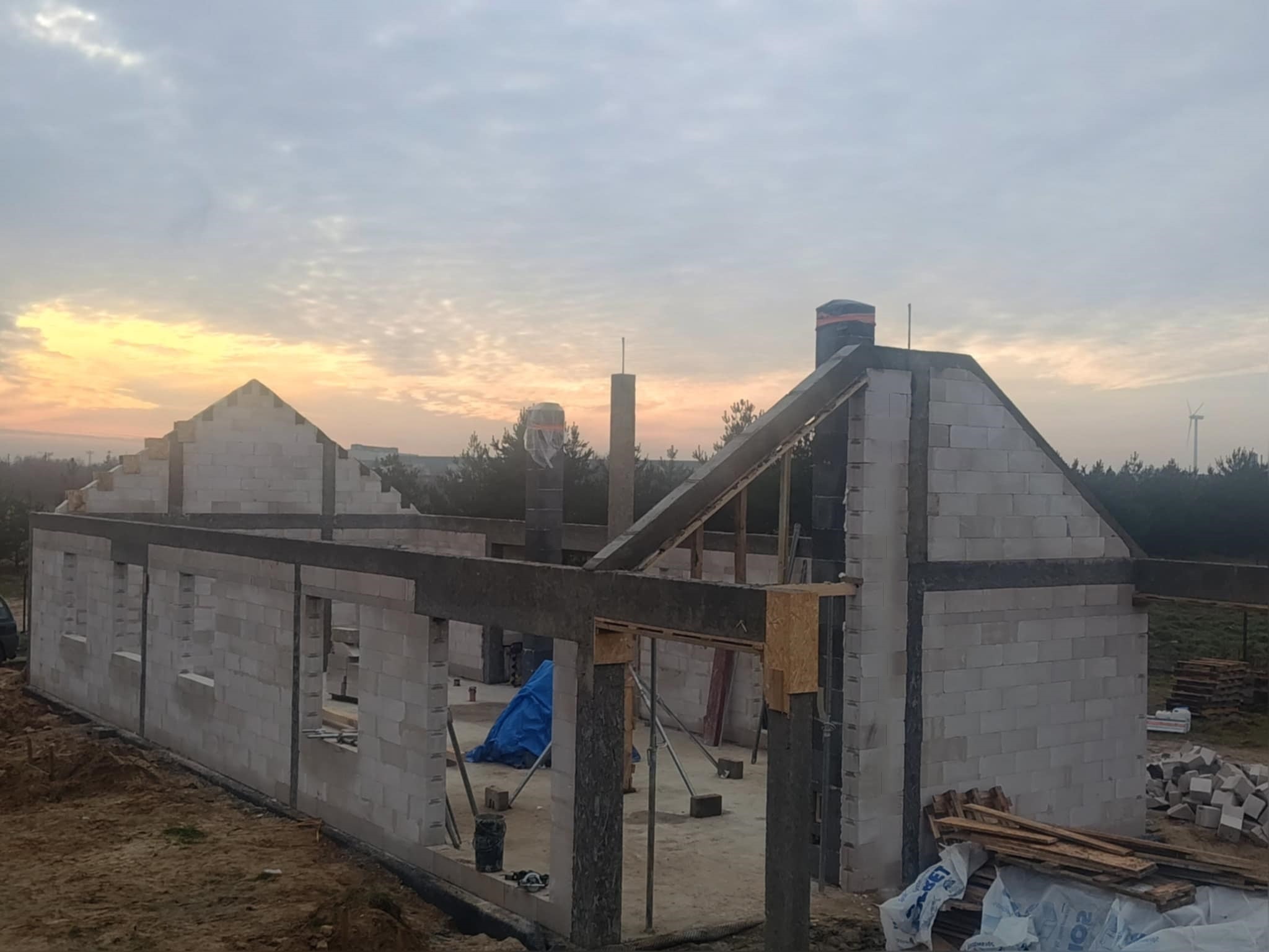 Budowa domu jednorodzinnego w stanie surowym otwartym, ściany z bloczków, widoczne elementy konstrukcyjne dachu, w tle zachodzące słońce i turbina wiatrowa.