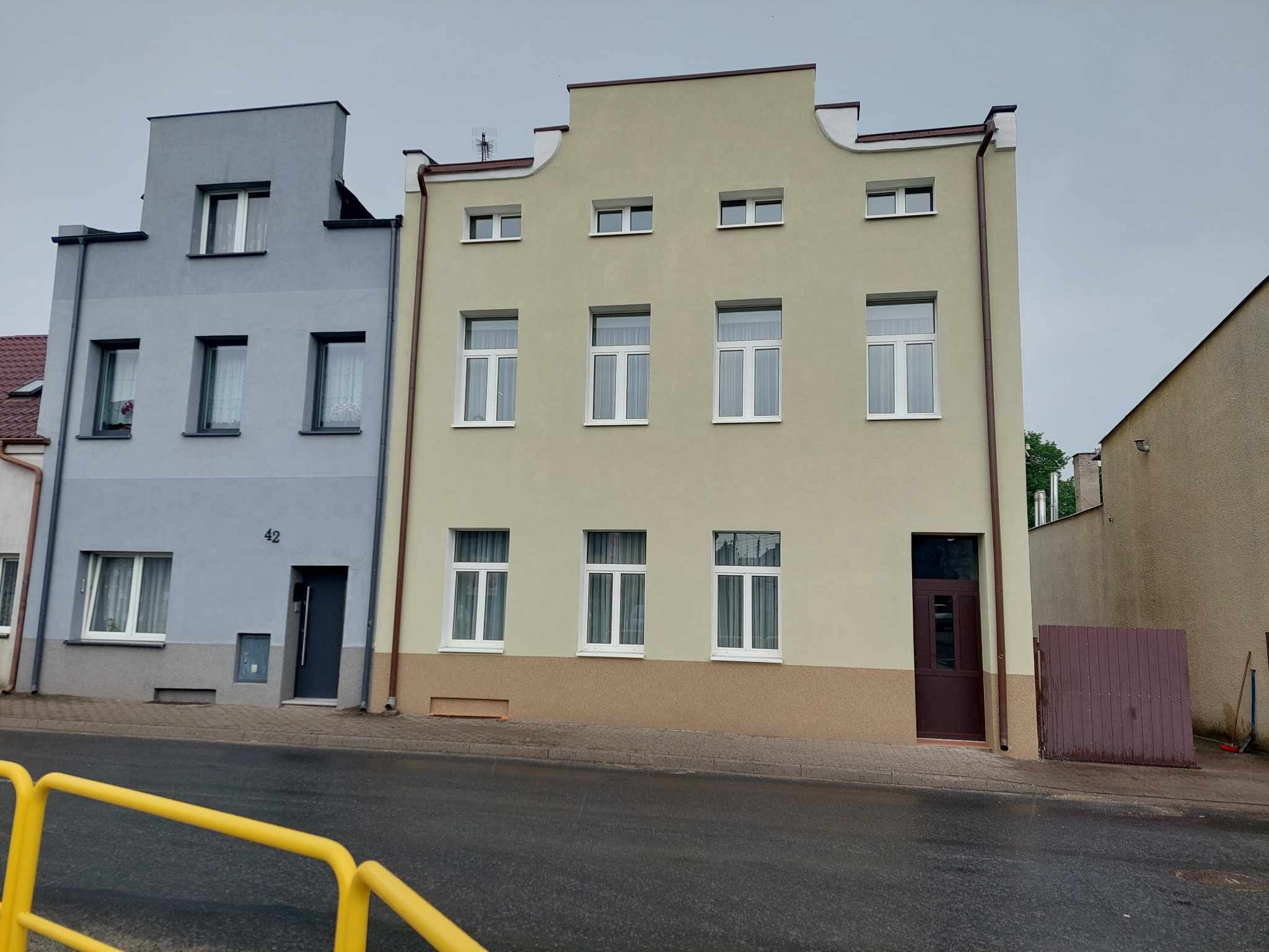 Odnowiona fasada dwukondygnacyjnego budynku z beżową elewacją i białymi oknami, sąsiadującego z szarym budynkiem z numerem 42. Widoczne obróbki blacharskie.