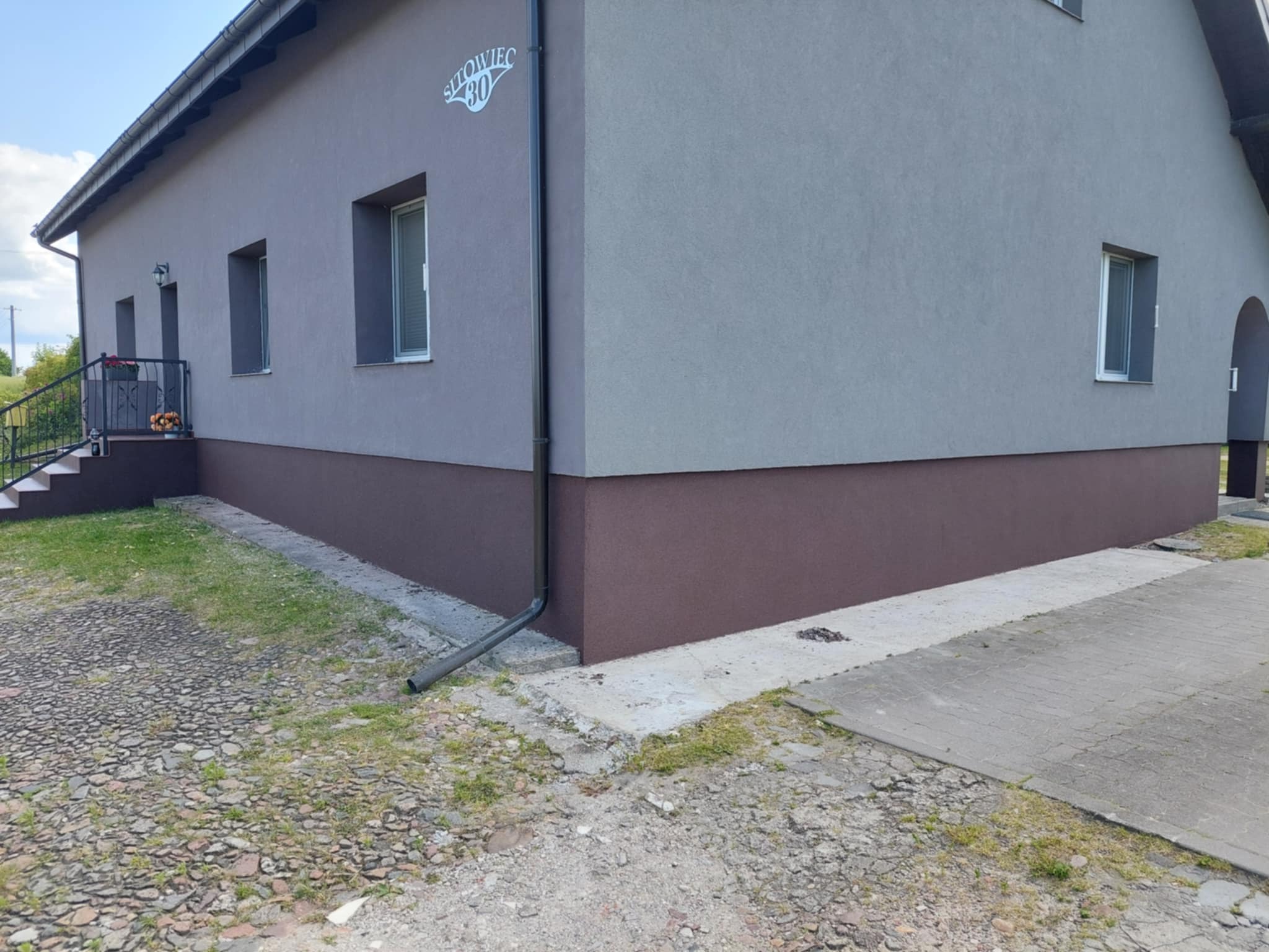 Elewacja domu jednorodzinnego z szarą górną częścią i brązowym cokołem, widoczny numer domu na rogu budynku oraz fragment chodnika i trawnika.