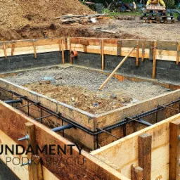 Szkielet ławy fundamentowej z drewnianym szalunkiem i zbrojeniem stalowym, widoczny wykop ziemny i bloczki betonowe, na dalszym planie sterta ziemi i materiały budowlane.