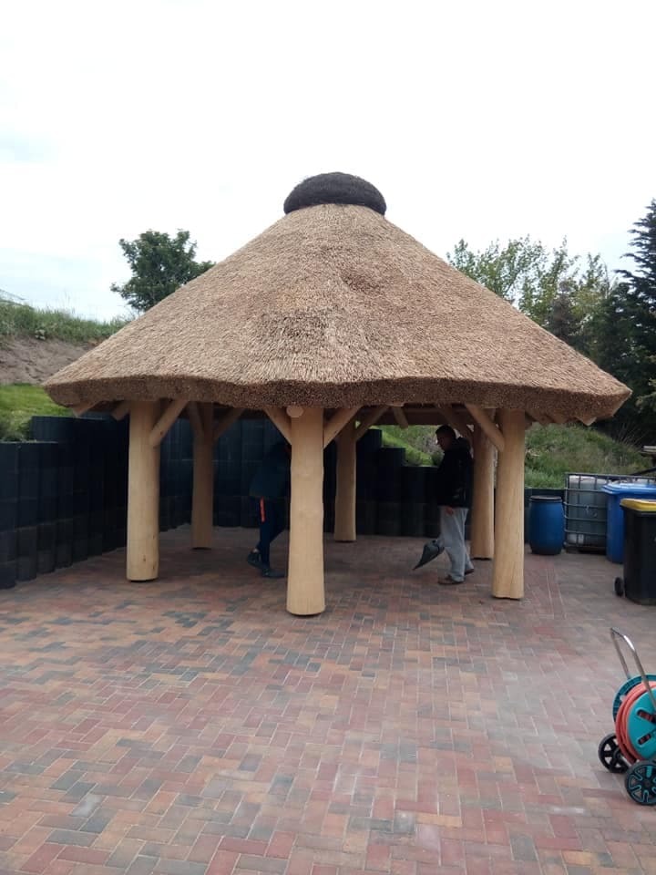 Altana z drewnianą konstrukcją i strzechą, na tle zieleni i kostki brukowej, z widocznymi postaciami wewnątrz.