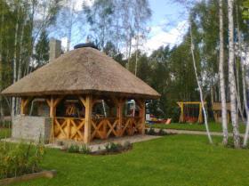 Drewniana altana z trzcinowym dachem w Trzebnicy, otoczona zielonym trawnikiem i brzozowym lasem. Widoczny fragment murowanego grilla i huśtawka w tle.