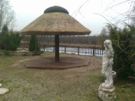 Altana kryta strzechą z ciemnym, drewnianym słupem, na ośmiokątnej, betonowej podstawie, z rzeźbą kobiety w ogrodzie, w pochmurny dzień.