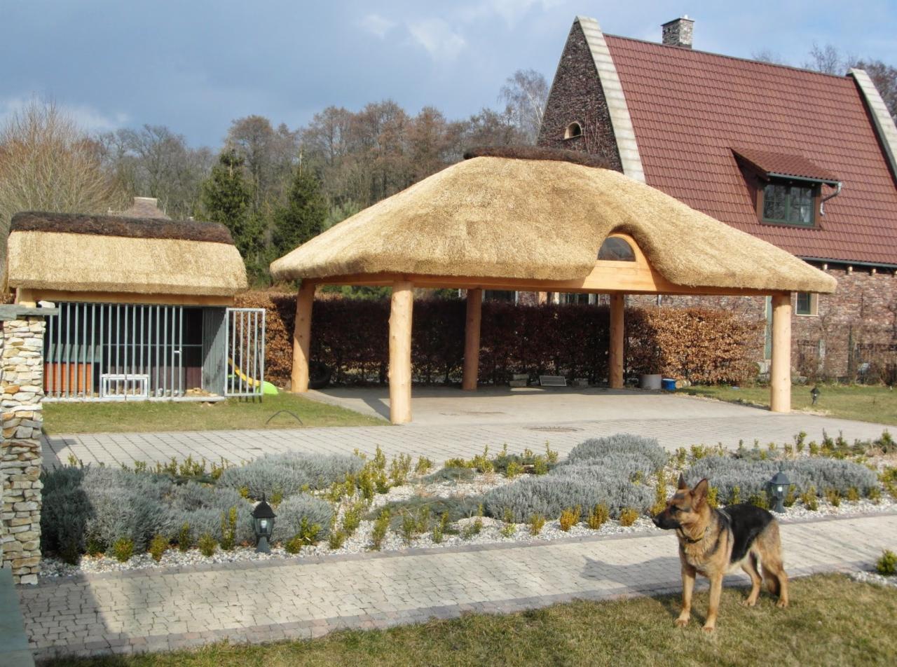 Zadaszenie z trzciny i domek dla psa z trzcinowym dachem, otoczone zielenią i brukowanym podjazdem. Owczarek niemiecki stoi na trawniku. W tle dom z czerwoną dachówką.
