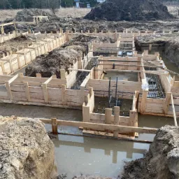 Zalane wodą szalunki fundamentowe zbrojone stalą, widoczne wykopy ziemne i pryzma ziemi w tle.