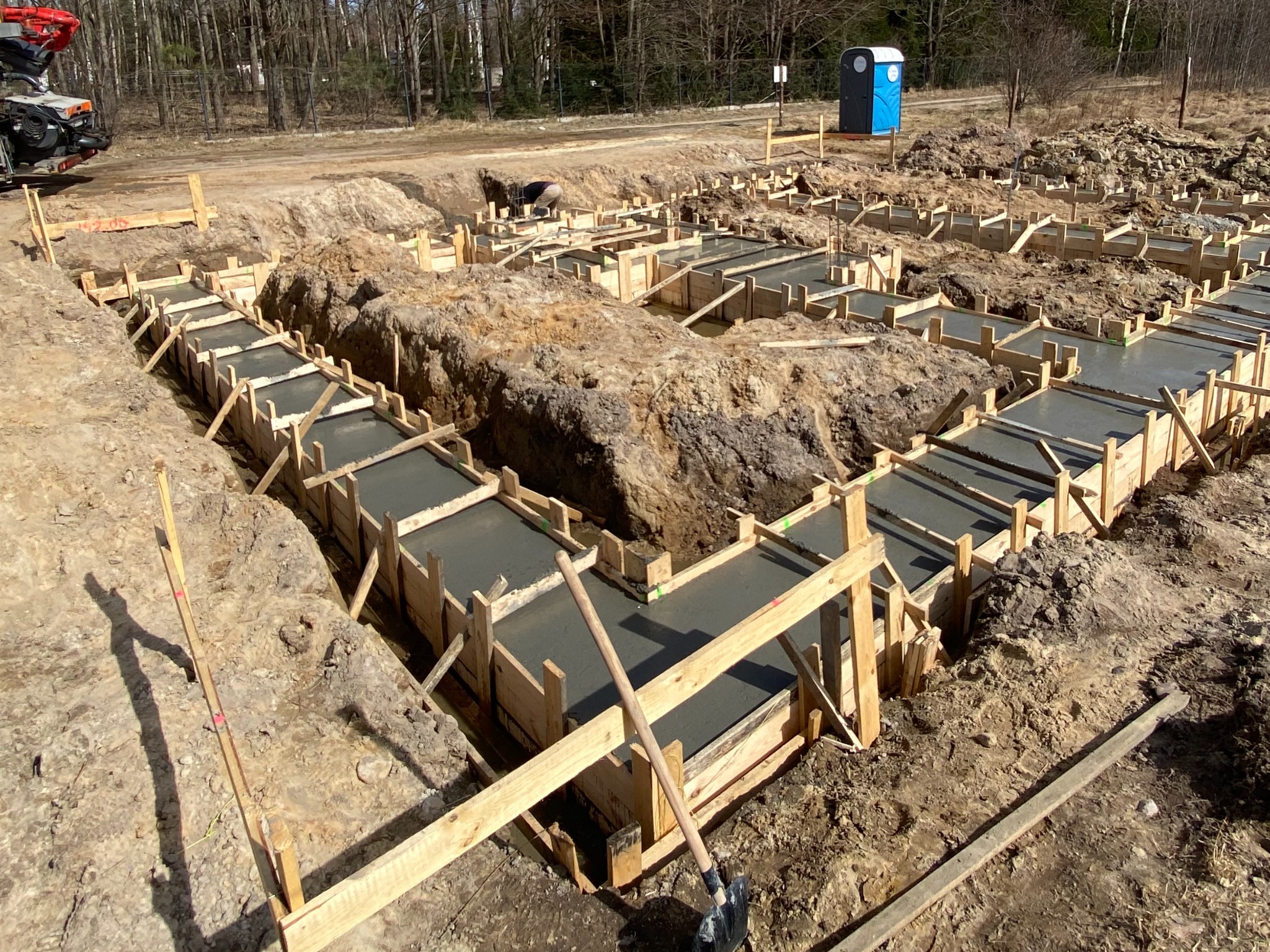 Wylane betonowe fundamenty w drewnianym szalunku, widoczny fragment betonomieszarki i przenośna toaleta w tle.