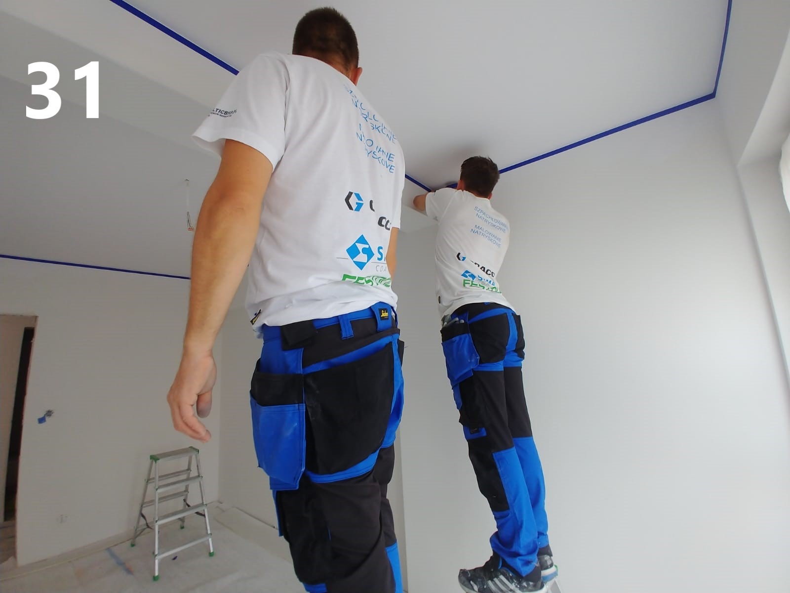 Dwóch pracowników w firmowych koszulkach, na drabinach, zabezpiecza taśmą malarską krawędzie sufitu w narożniku pomieszczenia przed rozpoczęciem prac wykończeniowych.
