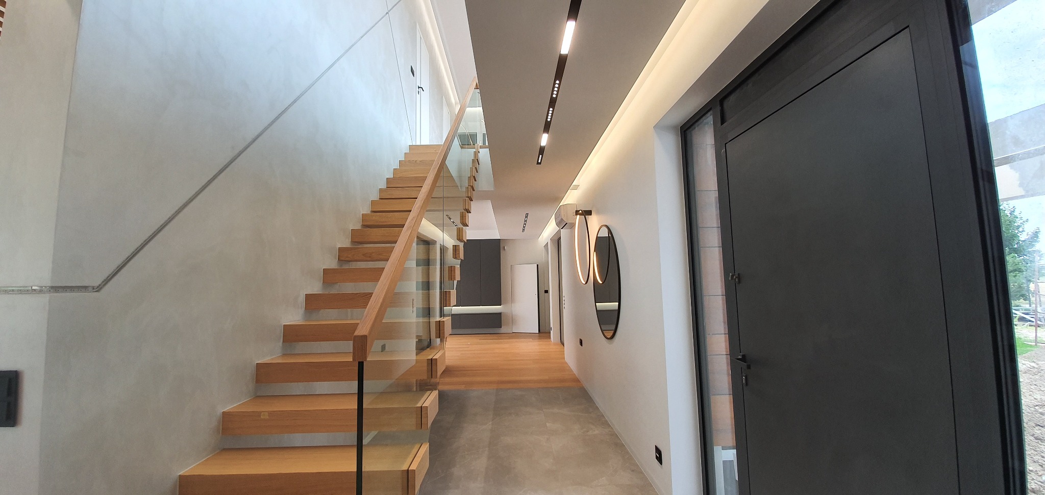 Nowoczesne wnętrze domu z drewnianymi schodami ze szklaną balustradą i gładką, szarą ścianą, oświetlone listwami LED na suficie.