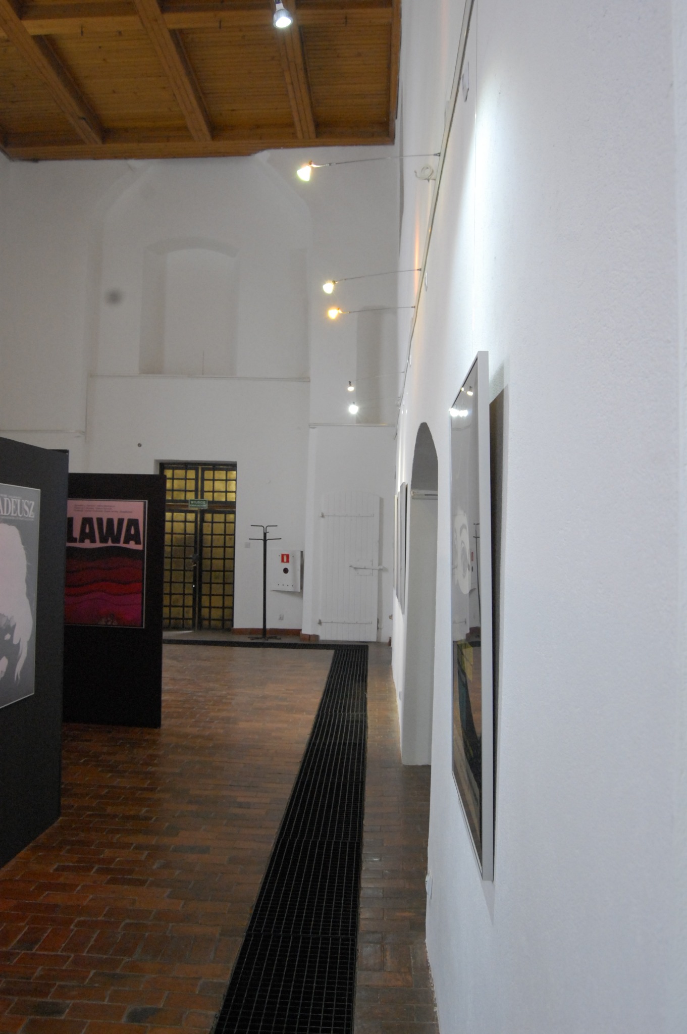 Wnętrze galerii z ceglaną podłogą, białymi ścianami oświetlonymi punktowymi lampami, z widocznymi plakatami i obrazami na ścianach.