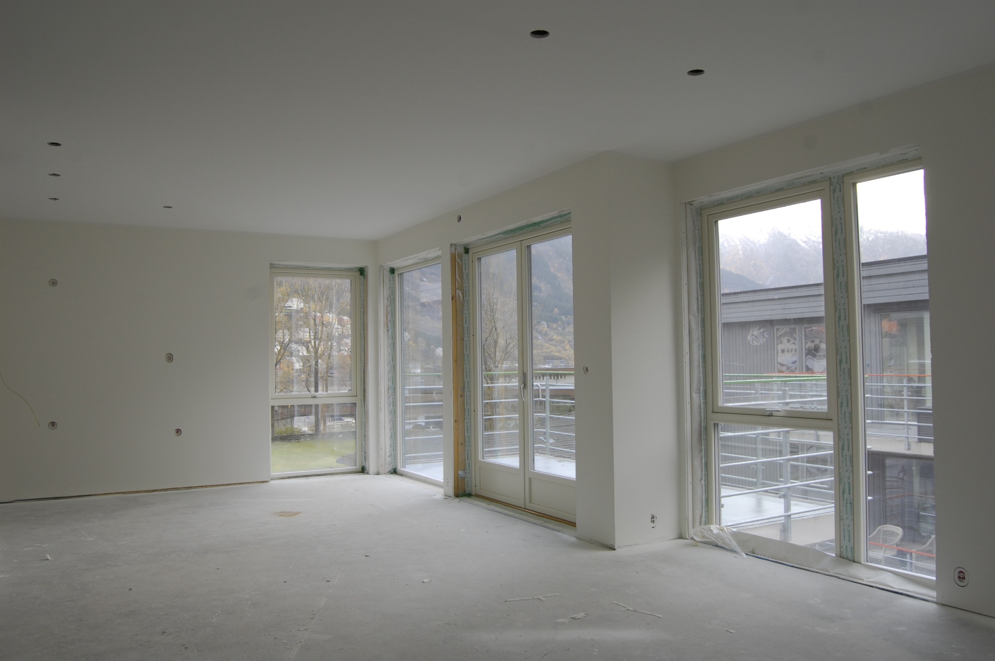 Surowe wnętrze z dużymi oknami i przygotowanymi ścianami do wykończenia, widoczne otwory na oświetlenie sufitowe i instalacje elektryczne w ścianach.