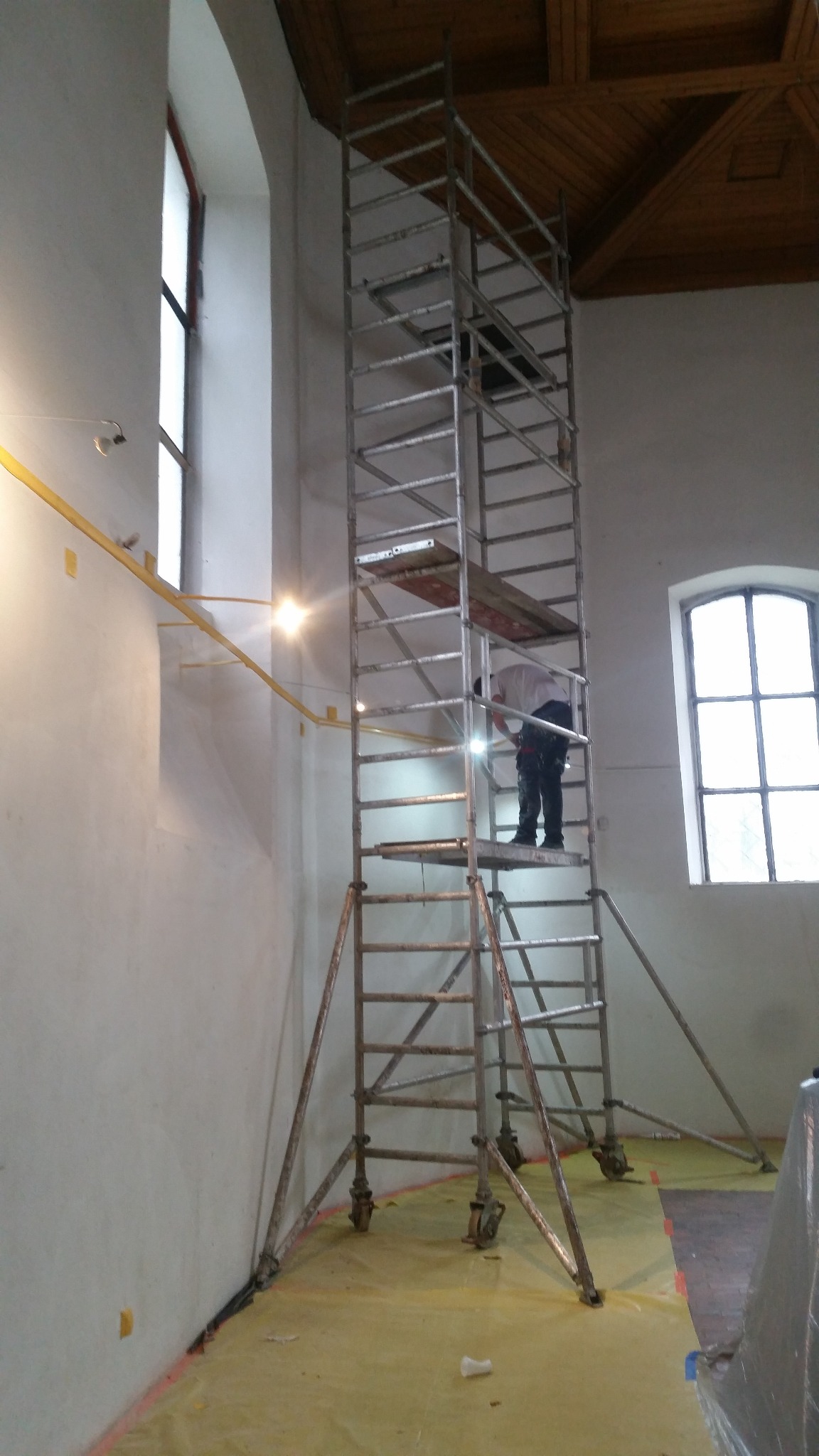 Pracownik na rusztowaniu w wysokim wnętrzu z drewnianym stropem, przygotowujący ścianę do prac wykończeniowych, zabezpieczona folią podłoga.