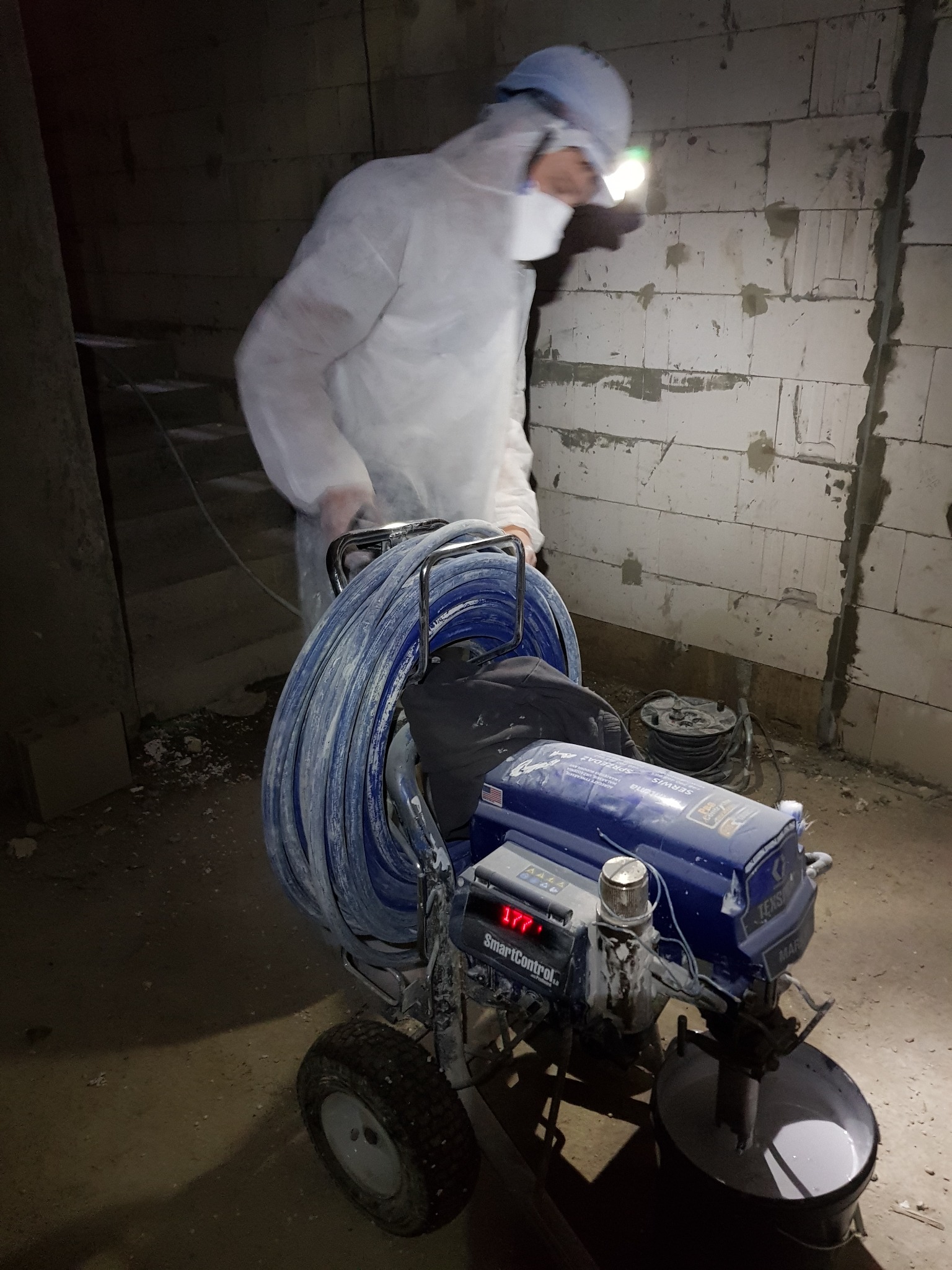 Pracownik w kombinezonie ochronnym, oświetlony latarką czołową, obsługuje agregat do natryskowego nakładania masy na ścianę z betonu komórkowego; widoczne ślady wcześniejszych prac.