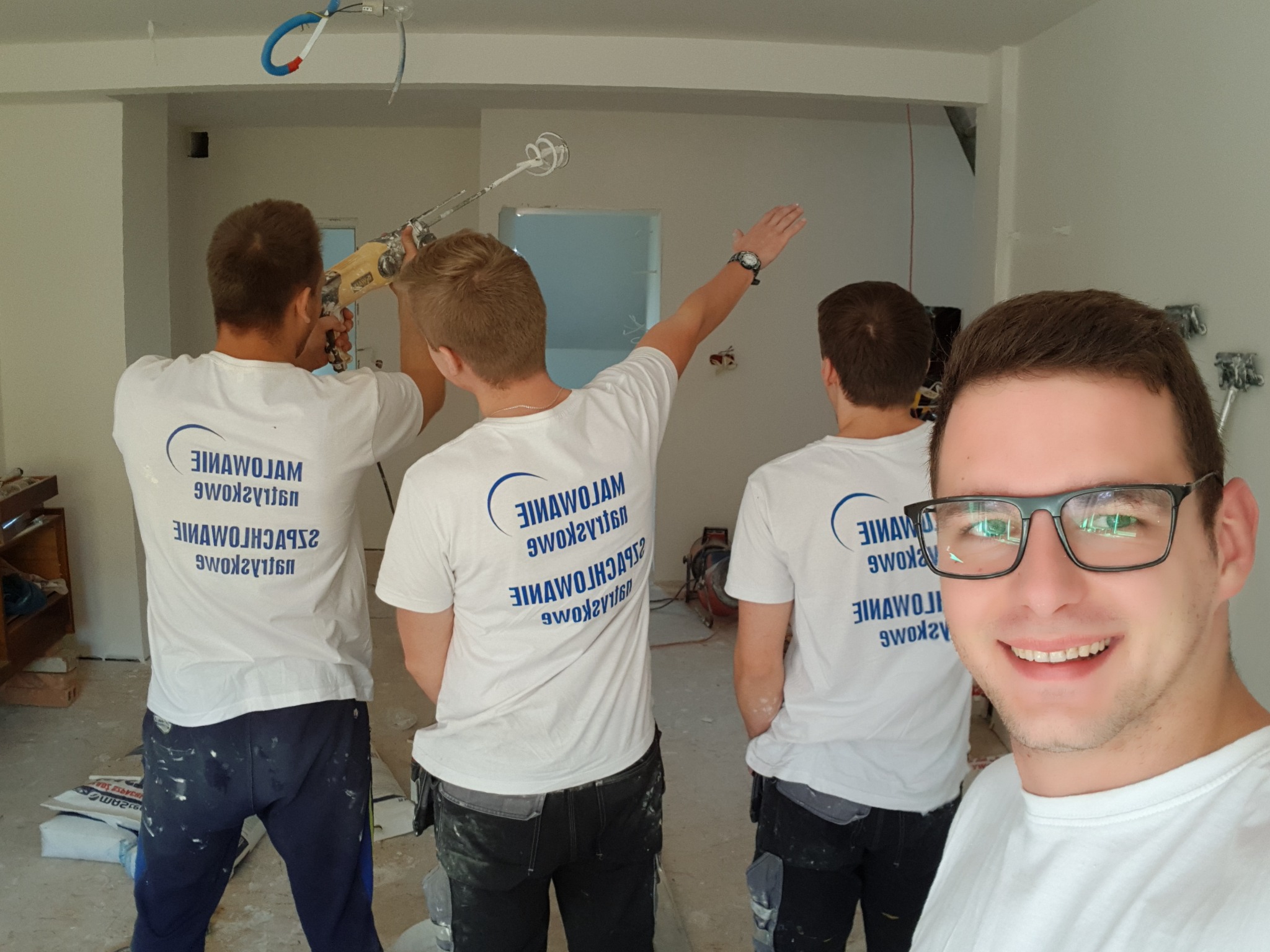 Ekipa remontowa w trakcie prac wykończeniowych, jeden z pracowników uśmiecha się do kamery podczas gdy pozostali mieszają zaprawę i wskazują miejsce do poprawki.