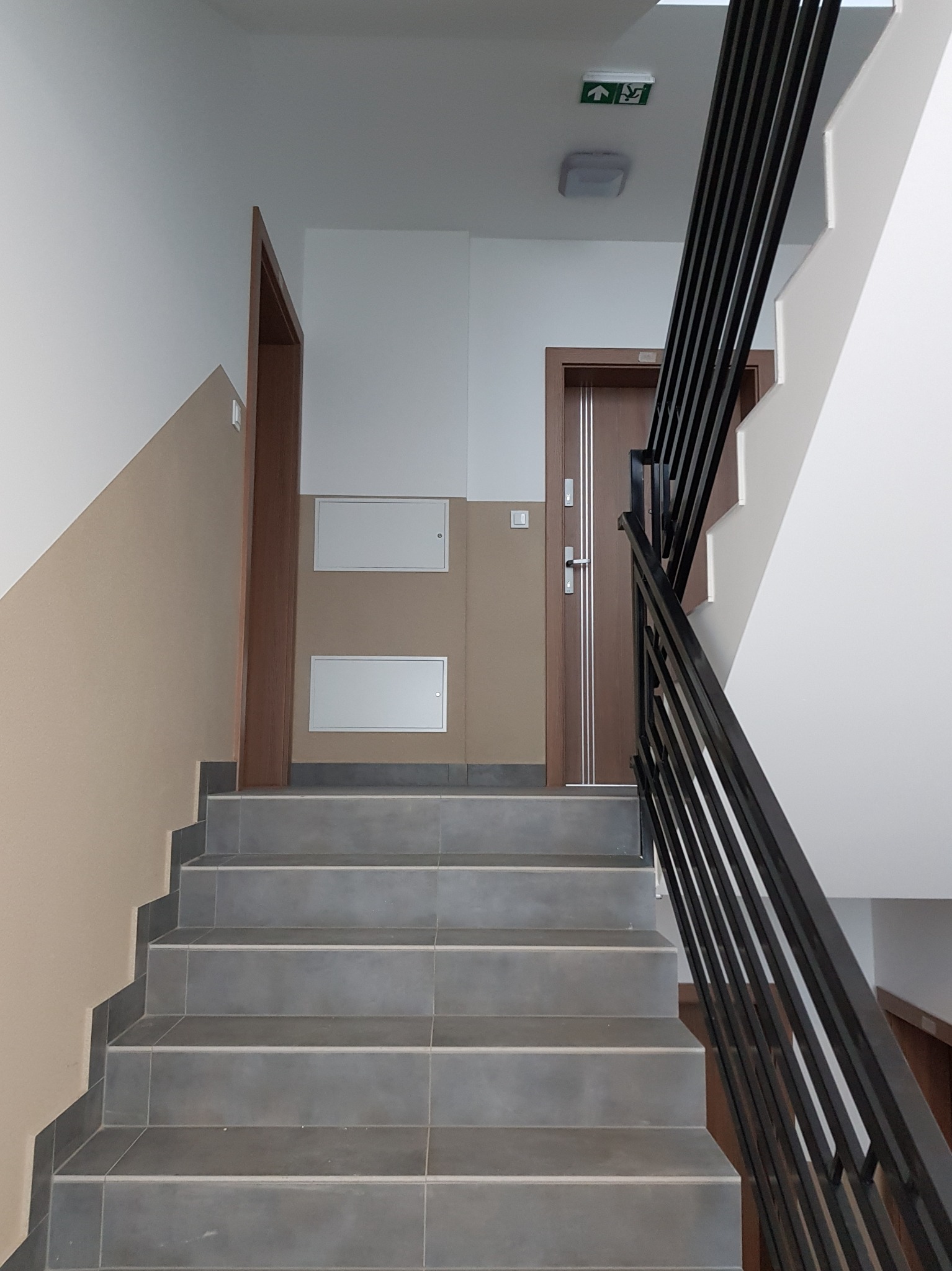Klatka schodowa w budynku biurowym z szarymi płytkami na schodach, brązowymi drzwiami i ścianami pomalowanymi w dwóch odcieniach beżu i bieli.