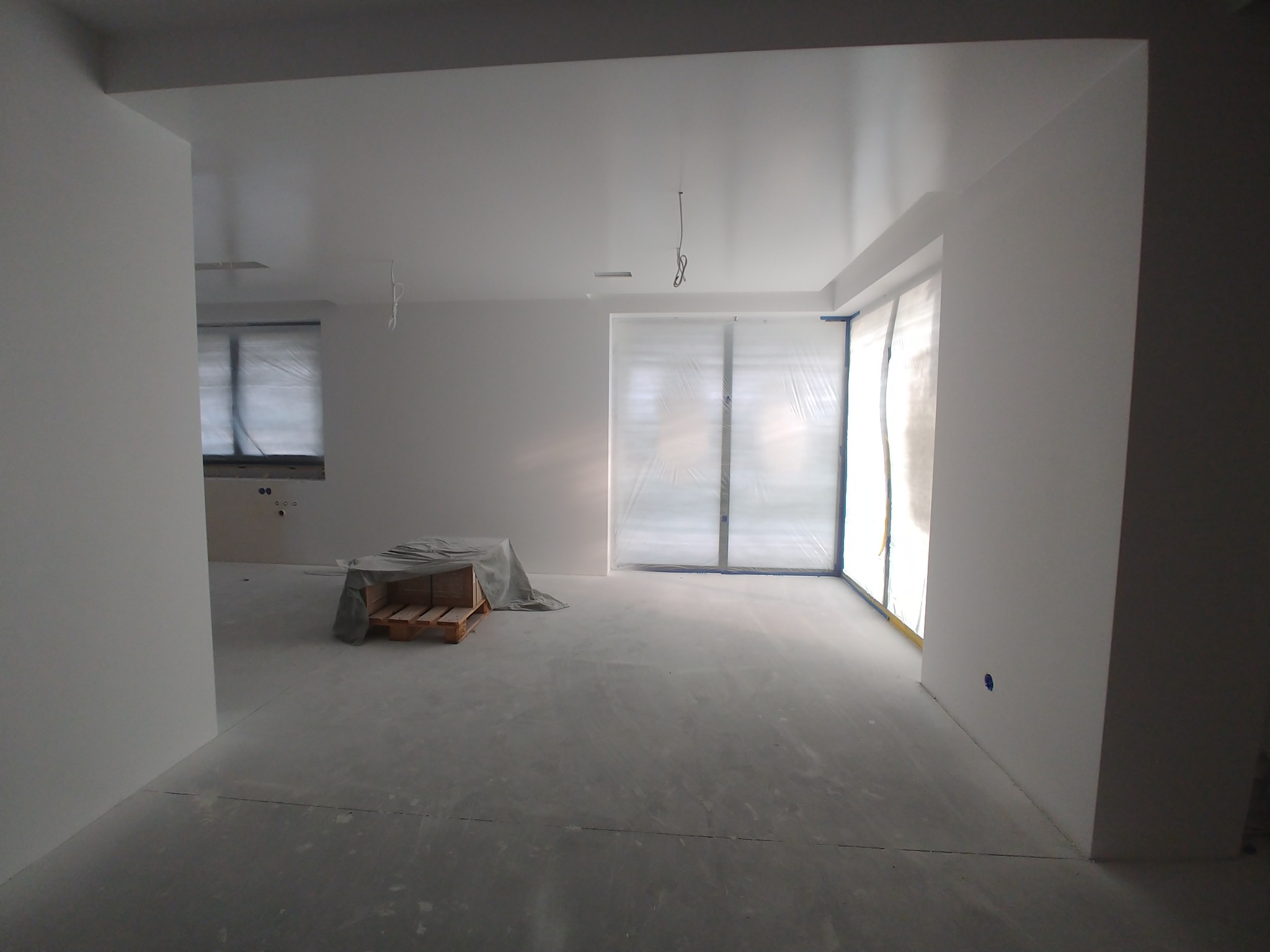 Wnętrze w trakcie prac wykończeniowych, ściany i sufit świeżo pokryte białą farbą, okna zabezpieczone folią malarską, podłoga zakurzona, w tle paleta przykryta folią.