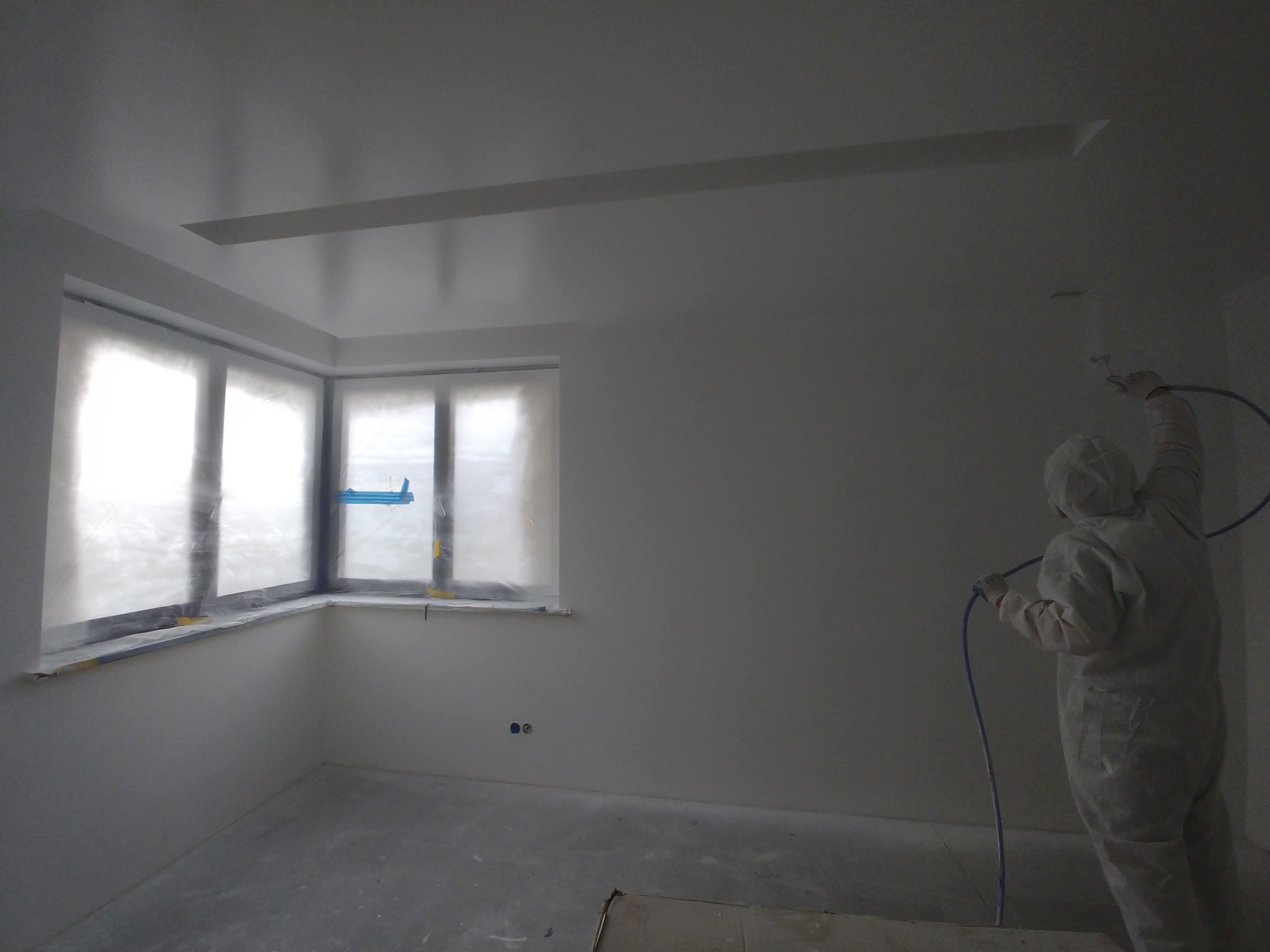 Pracownik w kombinezonie ochronnym natryskuje farbę na ścianę w pomieszczeniu z oknem zabezpieczonym folią malarską.