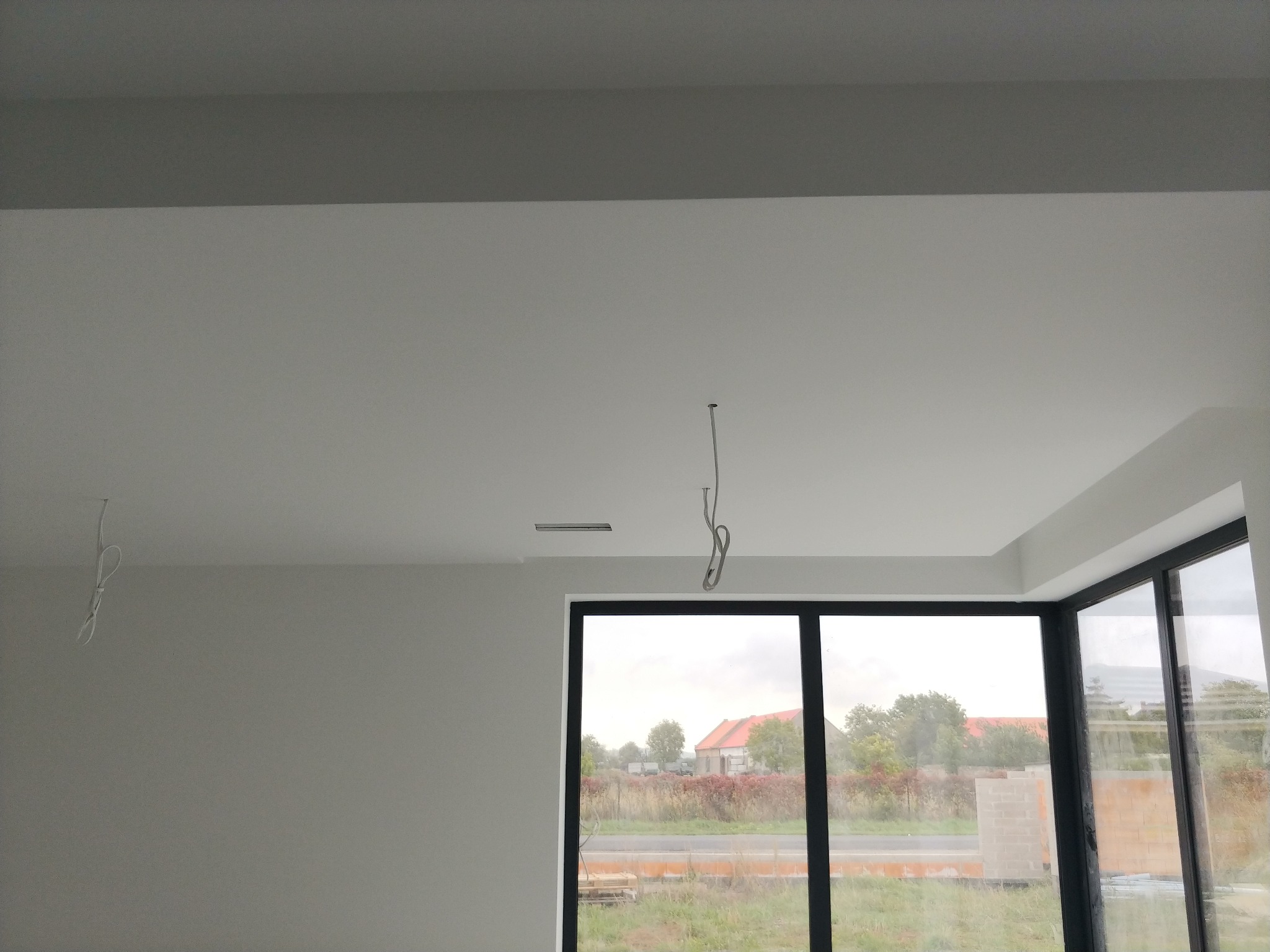 Wnętrze po odświeżeniu, ściany i sufit pomalowane na biało, widoczne wystające kable oświetleniowe i duże okno z widokiem na zewnątrz.