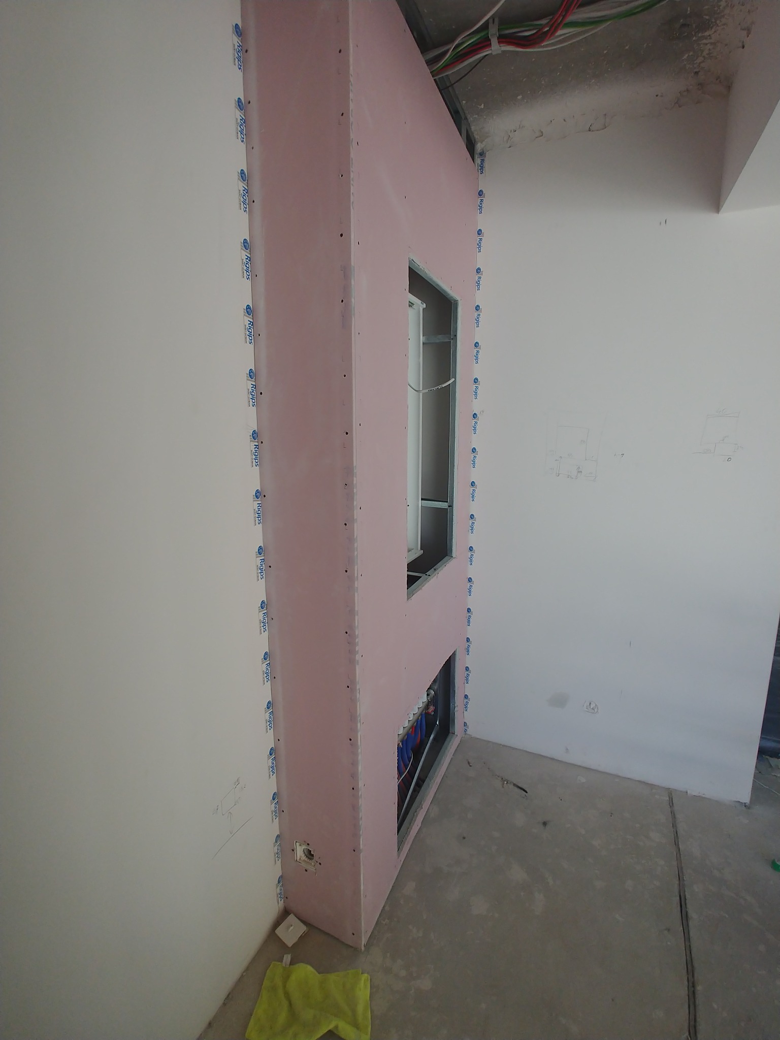 Montaż płyty gipsowo-kartonowej na stelażu metalowym z widocznymi instalacjami wewnątrz ściany, częściowo zakryte otwory, widoczne oznaczenia ołówkiem na sąsiedniej ścianie, podłoga w stanie...