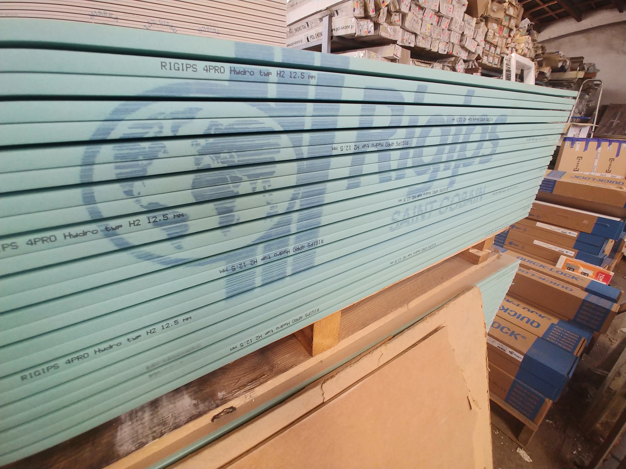 Stos zielonych płyt gipsowo-kartonowych Rigips 4PRO Hydro typu H2 12.5 mm, ułożonych na drewnianych paletach w magazynie budowlanym, z widocznymi kartonami Quick-Lock w tle.